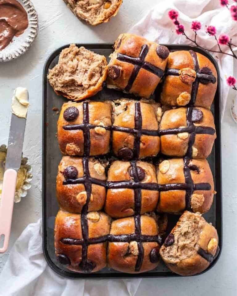 Chocolate hot cross buns on a tray with ingredients surrounding it.
