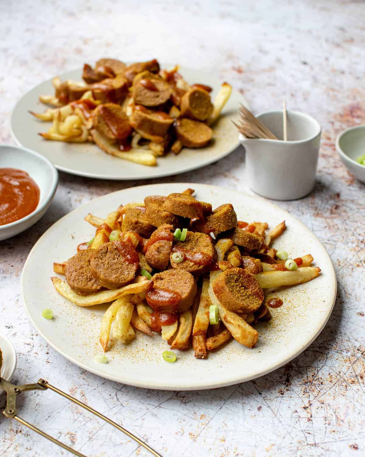Vegan currywurst with fries and curry ketchup on two plates and a pot of more sauce.