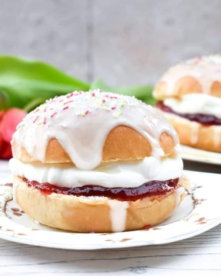 Iced bun on a plate with jam and cream, topped with icing and spinkles.