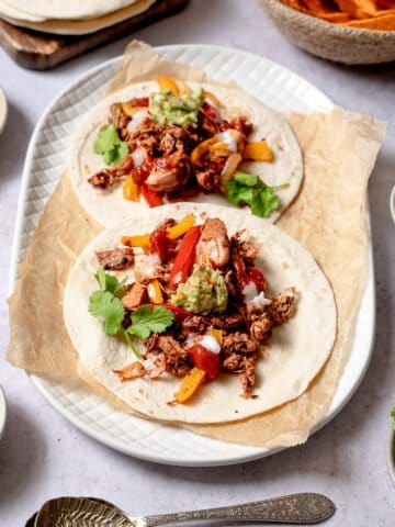 Two jackfruit fajitas on a plate with small bowls around it containing fresh coriander, avocado and salsa.