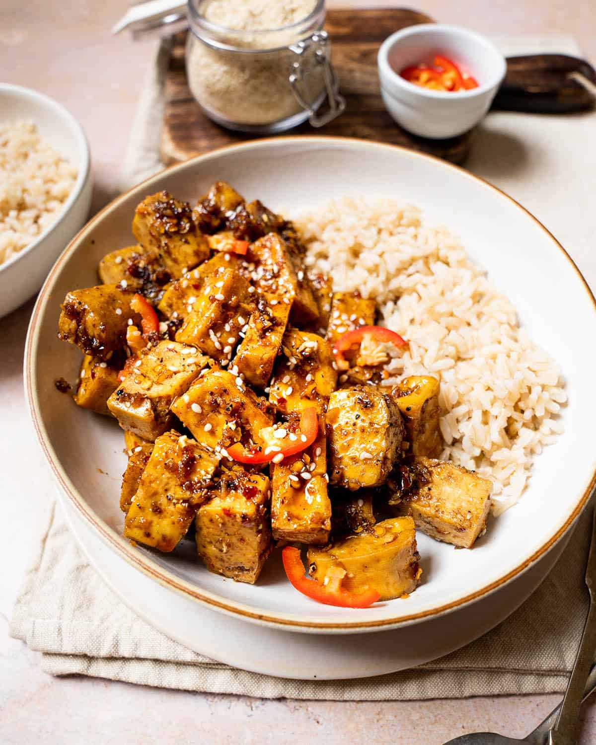 Pieces of Szechuan tofu in a bowl with brown rice and chillis and sesame seeds in the background.