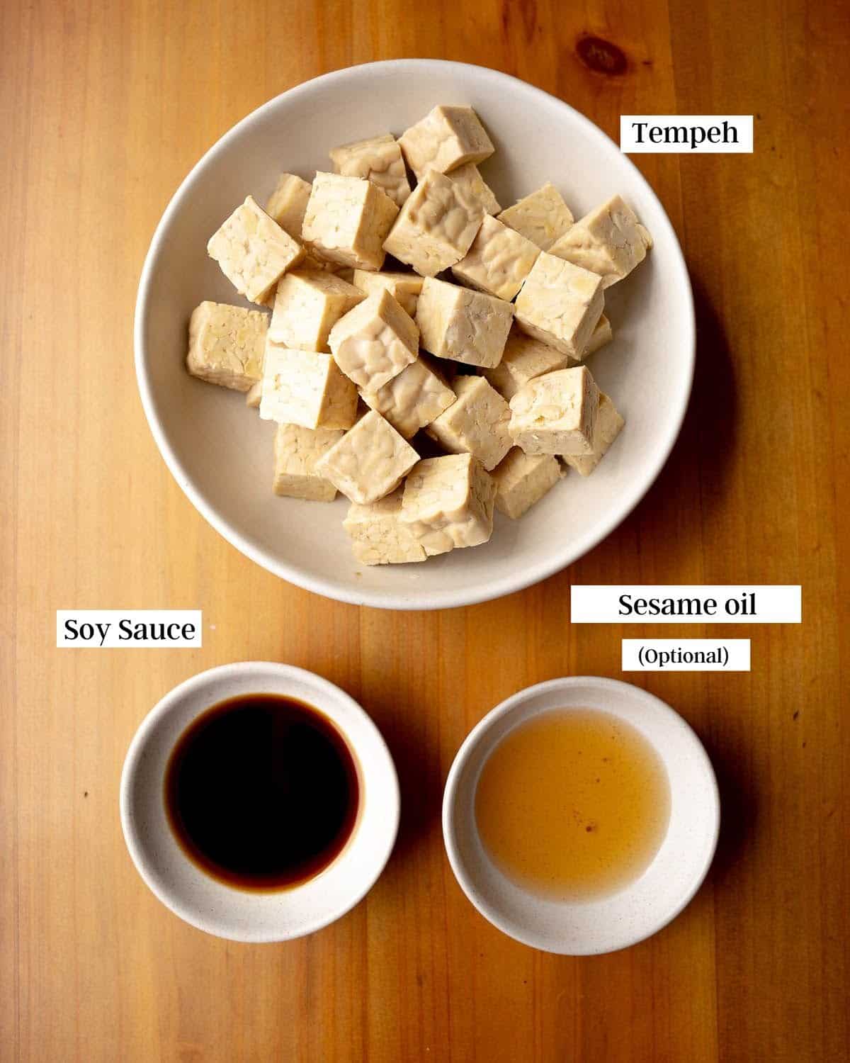 Tempeh chunks, soy sauce and oil in bowls on a wooden table.