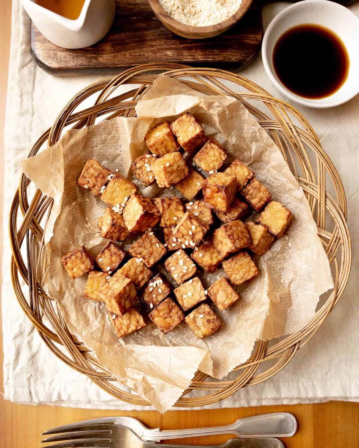 Tempeh chinks in a wicker basket with soy sauce and sesame seeds in small bowls.
