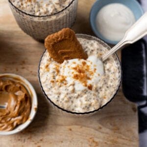 Two jars of Biscoff overnight oats topped with yoghurt and a biscuit. With a spoon.