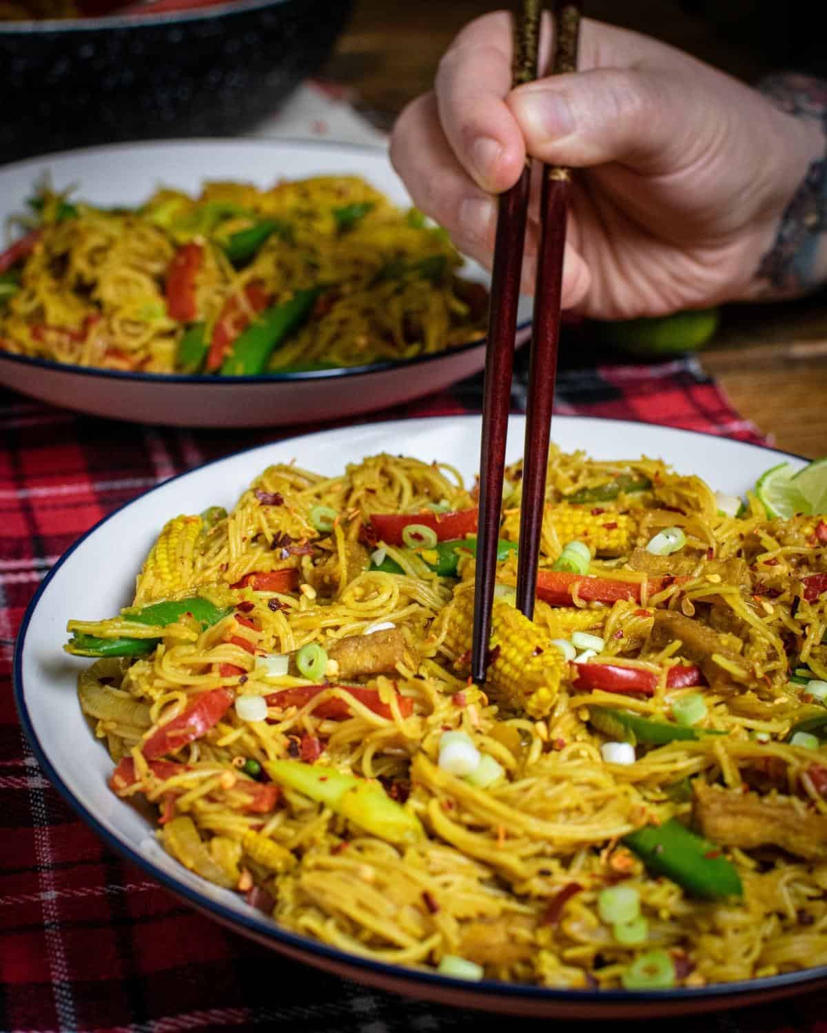 Tofu Singapore noodles on a plate with a hand holding chopsticks.
