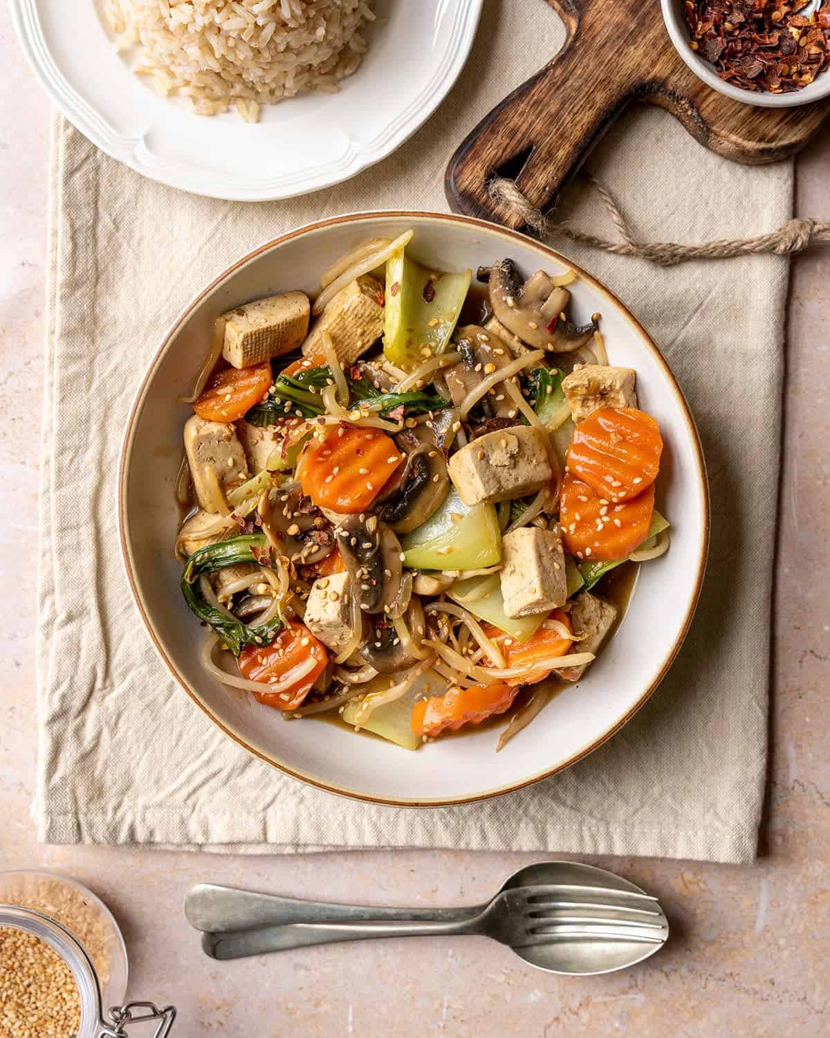 Vegan chop suey in a bowl with rice on the side and cutlery.