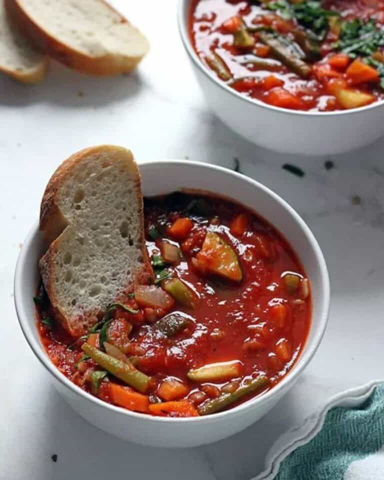 A bowl of minestrone soup with bread dunked into it.