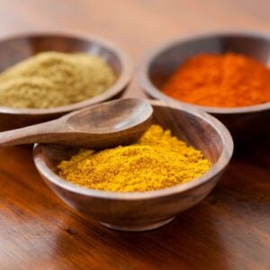 Cumin powder, turmeric powder and chili powder in wooden bowls with a wooden spoon.