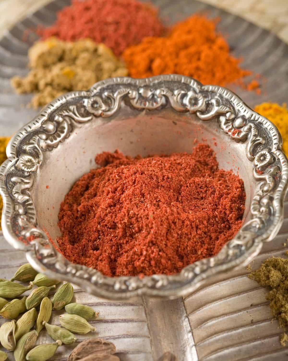Tandoor masala in a bowl with ground and whole spices surrounding it.