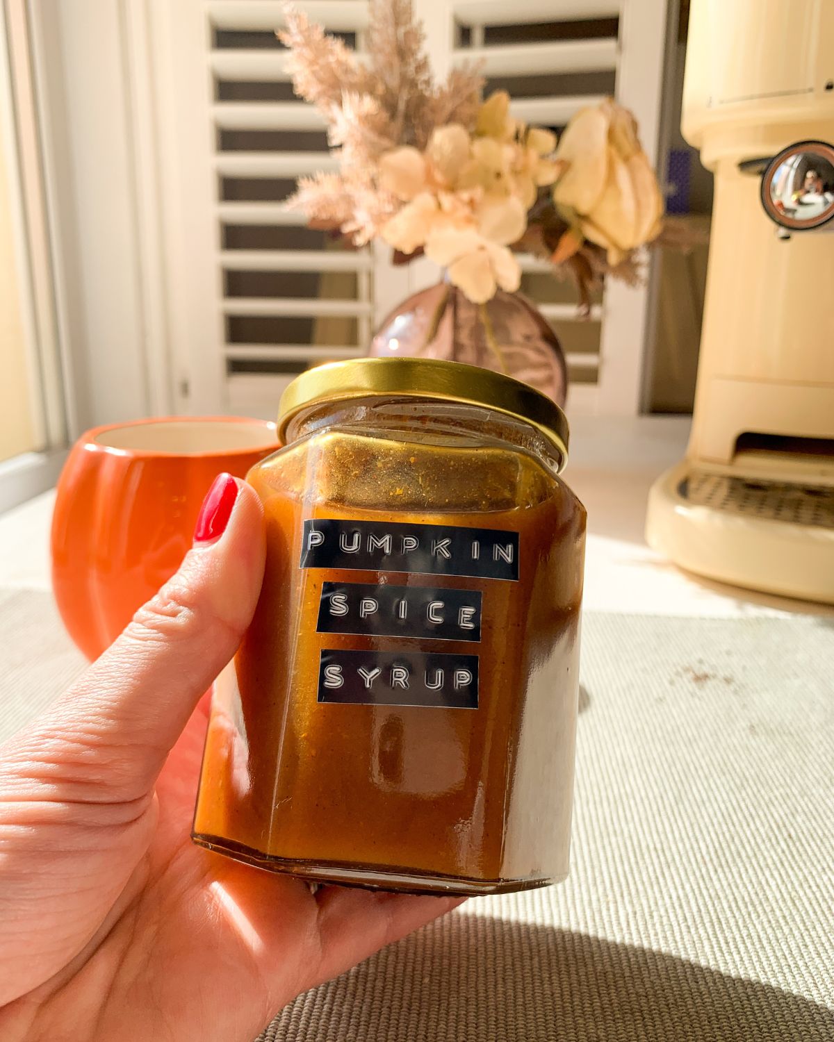 Pumpkin spice syrup in a jar with flowers and a coffee machine in the background.
